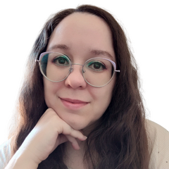 A pale person with long brown curly hair is wearing lilac cat-eye glasses and trans pride eye shadow while smiling gently toward the viewer and resting their chin on their hand